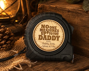 Puede incluir: Un flexómetro negro con una cara de madera grabada con las palabras "NO ONE MEASURES UP TO YOU DADDY" y los nombres Thaleia, Amira, Adonai y Lily. El flexómetro está sobre una superficie de arpillera.