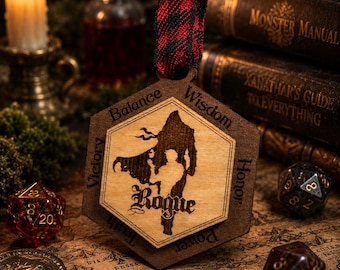 May include: Wooden hexagon-shaped ornament with a silhouette of a figure labeled "Rogue." The ornament has a red and black plaid ribbon. The words "Victory," "Balance," "Wisdom," "Honor," and "Power" are engraved around the edges. Dice and books are in the background.