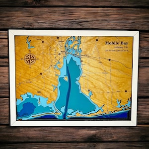 Puede incluir: Un mapa de madera enmarcado de Mobile Bay, Alabama, EE. UU., con un borde blanco. El mapa presenta una representación detallada de la bahía en tonos azules, con la tierra circundante en color marrón claro. El texto "Mobile Bay" y las coordenadas son visibles.