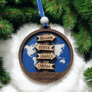 May include: A wooden Christmas ornament with a world map design and arrows pointing to Belgium, Kenya, Jamaica, and Canada. The ornament has a blue background and the text "Our Grand Adventures". It is hanging from a blue ribbon.