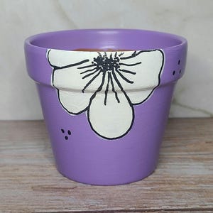May include: A hand-painted, small, purple flower pot. The pot features a large white flower design with black detailing and small black dots. The pot is made of ceramic and is suitable for small plants or succulents.