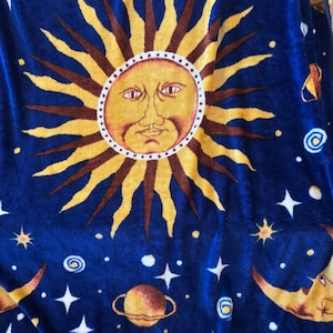 May include: A plush blue blanket featuring a large central sun with a human face, surrounded by yellow and brown rays. The design includes various planets, including one with rings, and white four-pointed stars scattered across the dark blue fabric.