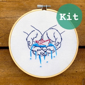 May include: Embroidery kit featuring a design of hands cradling a paper boat, with blue water cascading down. The boat is red and white. The word "Kit" is in a green circle. The design is on white fabric, set in a wooden embroidery hoop.