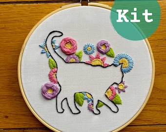 Pode incluir: Kit de bordado com a silhueta de um gato adornada com flores e folhas bordadas em cores vibrantes. O design inclui detalhes florais rosa, roxos, amarelos e azuis. A palavra "Kit" está em um círculo verde.