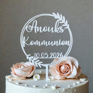 Könnte beinhalten: Ein weißer Tortenaufsatz mit den Worten "Anouks Kommunion" und dem Datum "10.05.2026" in einem kreisförmigen Design. Der Aufsatz ist mit floralen Akzenten verziert und sitzt auf einer Torte mit pfirsichfarbenen Rosen.