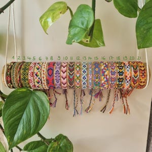 Può includere: Esposizione di 21 braccialetti dell'amicizia colorati, ognuno con motivi intrecciati unici e estremità a nappe. I braccialetti sono disposti su un supporto cilindrico in legno sospeso da cordoncini bianchi, con i numeri 1-21 sopra.