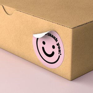 Puede incluir: Una pegatina circular rosa con una carita sonriente negra y el texto "&iexcl;gracias!" en una caja de cart&oacute;n marr&oacute;n.