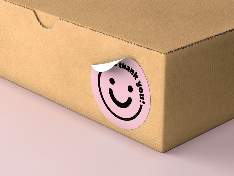 Puede incluir: Una pegatina circular rosa con una carita sonriente negra y el texto "&iexcl;gracias!" en una caja de cart&oacute;n marr&oacute;n.