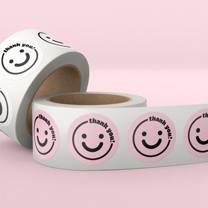 Puede incluir: Dos rollos de papel adhesivo blanco con pegatinas rosas y negras de "&iexcl;gracias!" con un dise&ntilde;o de cara sonriente.