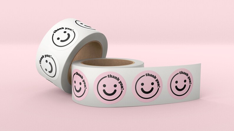Puede incluir: Dos rollos de papel adhesivo blanco con pegatinas rosas y negras de "&iexcl;gracias!" con un dise&ntilde;o de cara sonriente.