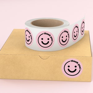 Puede incluir: Rollo de pegatinas de agradecimiento rosas y blancas con un dise&ntilde;o de cara sonriente. Las pegatinas est&aacute;n en una caja de cart&oacute;n marr&oacute;n.