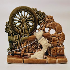 Può includere: Statuetta in ceramica raffigurante un cane incatenato a un filatoio. La statuetta è principalmente marrone e color crema, con un design dettagliato di un cane, una ruota e un fogliame. La base ha le lettere "R S".