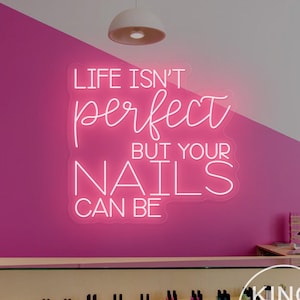 Op de afbeelding: Een neonbord met de tekst "LIFE ISN'T PERFECT BUT YOUR NAILS CAN BE" in een sierlijk lettertype, tegen een roze muur. Het bord bevindt zich in een nagelsalon, met nagellakflesjes tentoongesteld.