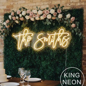 May include: A wedding reception scene featuring a three-tiered white cake with pink floral accents, wine glasses, and plates on a wooden table. A neon sign with the words "the Smiths" is illuminated against a green hedge backdrop, adorned with roses and greenery.