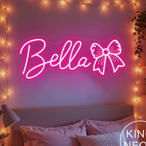 Op de afbeelding: Een roze neonbord met de naam "Bella" in cursief schrift en een strik. Het bord is aan een muur boven een bed bevestigd. Het bord is van King Neon.