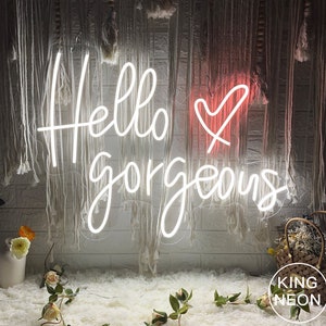 K&ouml;nnte beinhalten: Ein wei&szlig;es Neon-Schild mit dem Text "Hello gorgeous" und einem rosa Herzen. Das Schild h&auml;ngt an einer Wand mit einem wei&szlig;en und grauen, strukturierten Hintergrund.