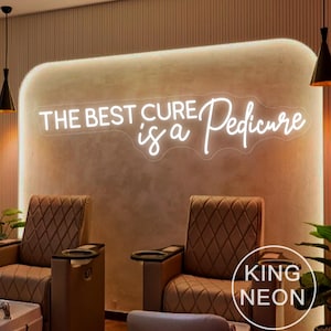 Könnte beinhalten: Ein Neon-Schild mit dem Text "The best cure is a Pedicure" in weißer, kursiv geschriebener Schrift. Das Schild ist an einer Wand mit hellbraunem Hintergrund angebracht. Im Vordergrund stehen zwei braune Ledersessel.