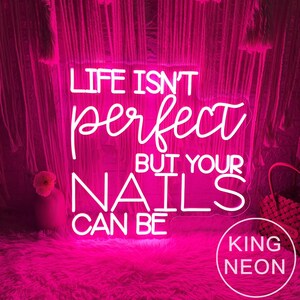 Op de afbeelding: Een helder roze neonbord met de tekst "LIFE ISN'T perfect BUT YOUR NAILS CAN BE". Het bord wordt omgeven door een roze, pluizig tapijt en een decoratieve achtergrond. Het bord heeft ook de woorden "KING NEON" in een cirkel.