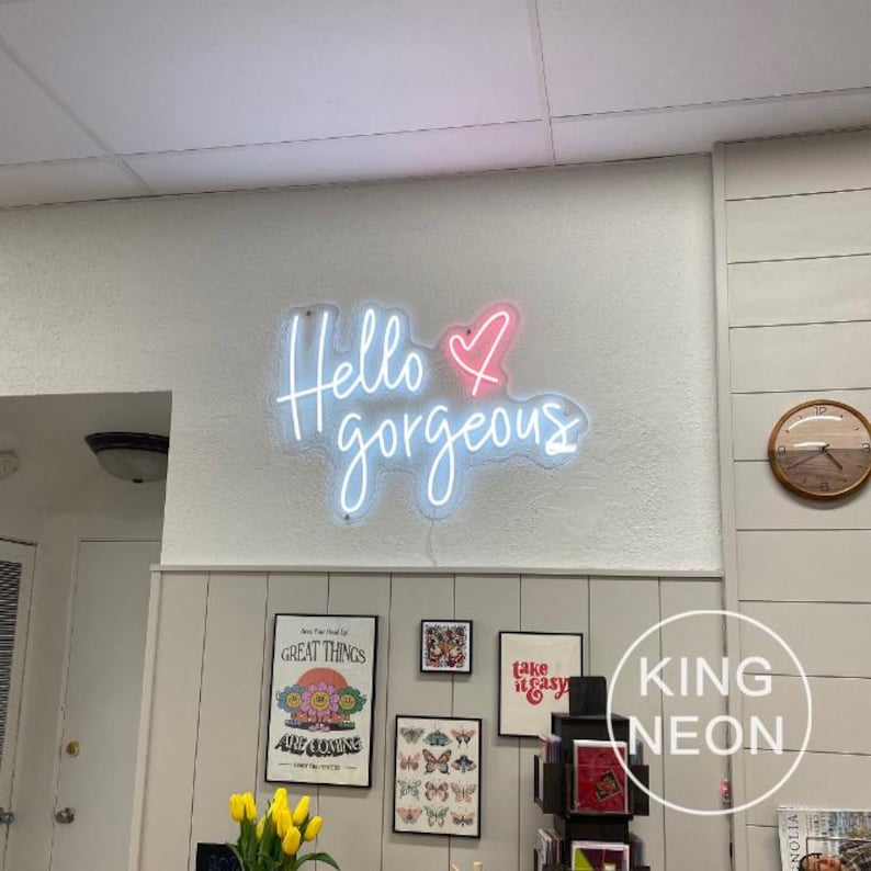K&ouml;nnte beinhalten: Neon-Schild mit dem Text "Hello gorgeous" in Wei&szlig; mit einem rosa Herzen. Das Schild ist an einer wei&szlig;en Wand mit verschiedenen gerahmten Kunstwerken und einer Uhr befestigt.