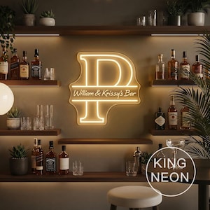 May include: A warm white neon sign featuring a large letter P with the text William & Krissy's Bar centered horizontally across the letter. The sign is mounted on a wall between wooden shelves stocked with various glass liquor bottles and glasses.