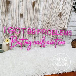 K&ouml;nnte beinhalten: Rosa Neon-Schild mit dem Text "I got 99 problems but my nails ain't one". Das Schild h&auml;ngt an einer wei&szlig;en Wand mit einem wei&szlig;en, flauschigen Teppich im Vordergrund. Der Text "KING NEON" ist in der rechten unteren Ecke sichtbar.