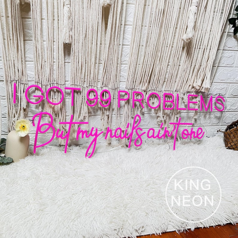 K&ouml;nnte beinhalten: Rosa Neon-Schild mit dem Text "I got 99 problems but my nails ain't one". Das Schild h&auml;ngt an einer wei&szlig;en Wand mit einem wei&szlig;en, flauschigen Teppich im Vordergrund. Der Text "KING NEON" ist in der rechten unteren Ecke sichtbar.