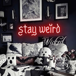 Op de afbeelding: Een rood neonbord met de tekst "stay weird" hangt aan een donkerblauwe muur. Het bord hangt boven een verzameling Halloween-thema kussens en decoraties, waaronder een spookvormig kussen en een kussen met een raaf.