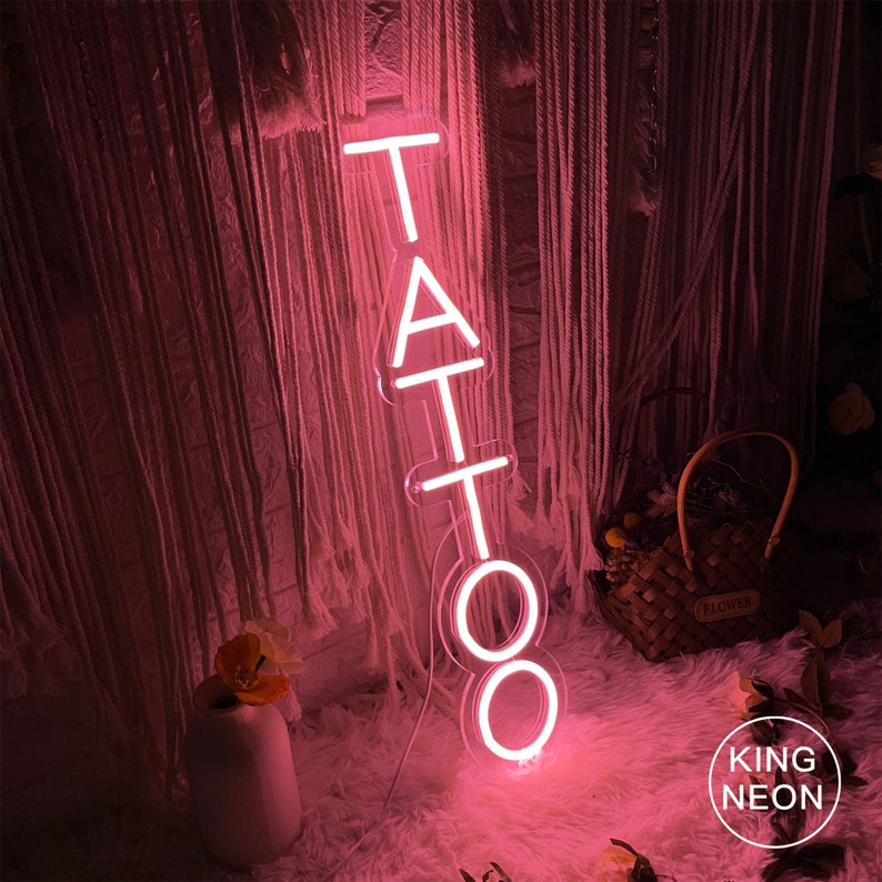 May include: Pink neon sign with the word "TATTOO" in white neon letters. The sign is lit up and casting a pink glow on the surrounding area. The sign is on a white fluffy surface. The text "KING NEON" is visible in the bottom right corner of the image.