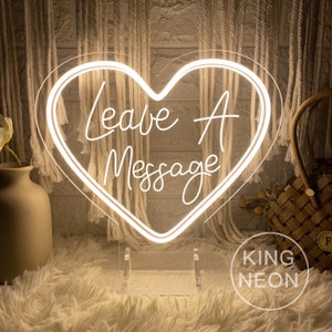 Könnte beinhalten: Ein weißes Neon-Schild in Herzform mit dem Text "Leave A Message" im Inneren. Das Schild steht auf einem klaren Acryl-Ständer. Das Schild befindet sich auf einer weißen, flauschigen Oberfläche.