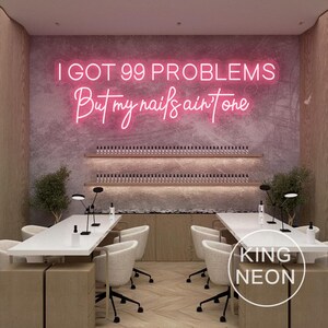 K&ouml;nnte beinhalten: Ein pinkfarbenes Neon-Schild mit dem Text "I got 99 problems but my nails ain't one". Das Schild h&auml;ngt an einer Wand in einem Nagelstudio. Im Vordergrund befinden sich zwei wei&szlig;e Tische mit St&uuml;hlen.