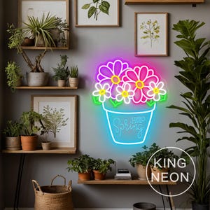 Può includere: Un'insegna al neon a forma di vaso di fiori con le parole "Hello Spring". I fiori sono rosa e viola con centri gialli e foglie verdi. L'insegna è circondata da piante e stampe botaniche incorniciate.