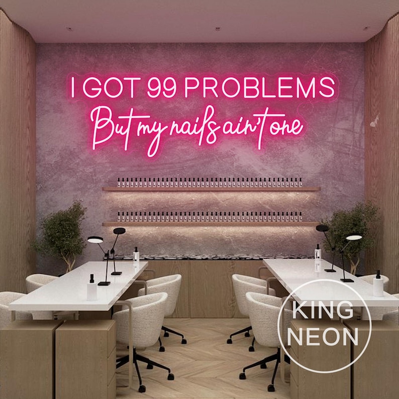 K&ouml;nnte beinhalten: Ein pinkfarbenes Neon-Schild mit dem Text "I got 99 problems but my nails ain't one" h&auml;ngt an einer Wand in einem Nagelstudio. Das Studio hat zwei Manik&uuml;re-Stationen mit wei&szlig;en Tischen und wei&szlig;en St&uuml;hlen. Das Studio hat auch eine Holzwand mit Regalen voller Nagellack.