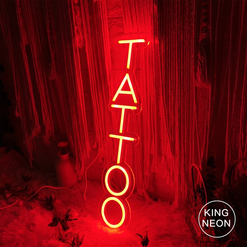 May include: A neon sign with the word "TATTOO" in red, lit up against a red background. The sign is made of individual letters, each with a white outline. The sign is lit up and casting a warm glow. The sign is from King Neon.