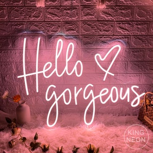 K&ouml;nnte beinhalten: Rosa Neon-Schild mit wei&szlig;er Aufschrift "Hello gorgeous" mit einem Herzsymbol zwischen den W&ouml;rtern. Das Schild befindet sich auf einer rosa flauschigen Oberfl&auml;che.