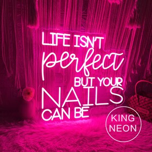 Op de afbeelding: Een helder roze neonbord met de tekst "LIFE ISN'T perfect BUT YOUR NAILS CAN BE". Het bord staat op een roze, pluizig oppervlak en heeft de tekst "KING NEON" in een cirkel.