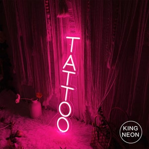 May include: Pink neon sign with the word "TATTOO" in white letters. The sign is hanging in front of a pink and white curtain.