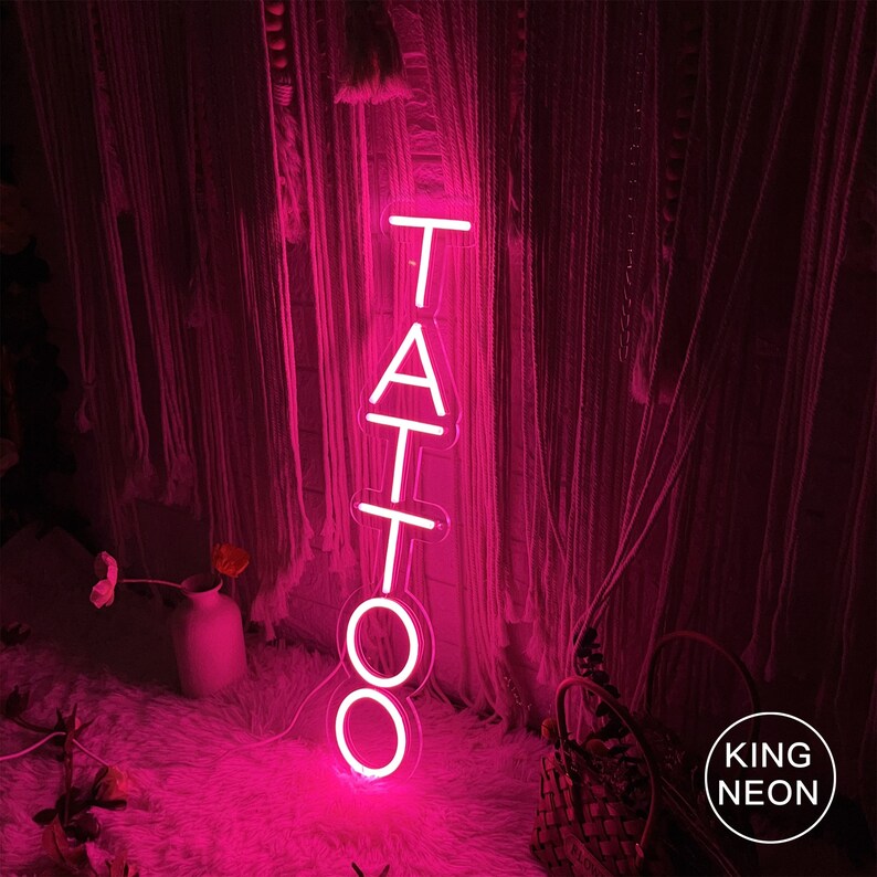 May include: Pink neon sign with the word "TATTOO" in white letters. The sign is hanging in front of a pink and white curtain.