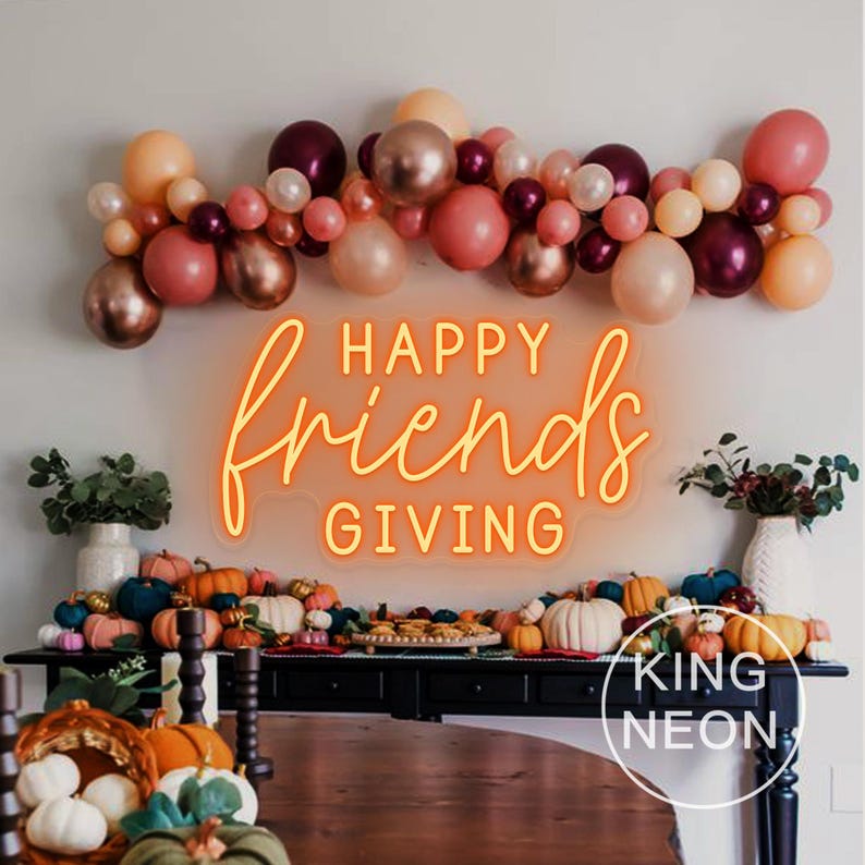 May include: A festive Thanksgiving display featuring a neon sign that reads "Happy Friends Giving." The sign is above a table decorated with pumpkins, gourds, and a balloon garland in shades of pink, peach, and burgundy.