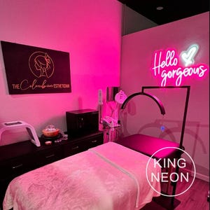 K&ouml;nnte beinhalten: Ein rosa Raum mit einer Massageliege, einem Neon-Schild mit der Aufschrift "Hello gorgeous" und einem Schild mit der Aufschrift "The Colombian Esthetician".