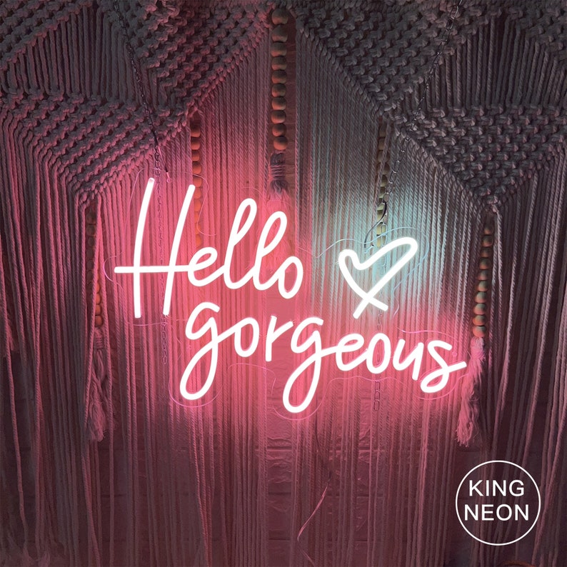 K&ouml;nnte beinhalten: Ein wei&szlig;es Neon-Schild mit dem Text "Hello gorgeous" und einem Herzsymbol. Das Schild h&auml;ngt an einer Wand mit einem rosa und wei&szlig;en Makramee-Wandbehang dahinter.