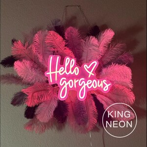 K&ouml;nnte beinhalten: Ein pinkfarbenes Neon-Schild mit dem Text "Hello gorgeous" mit einem Herzsymbol. Das Schild h&auml;ngt an einer Kette und ist von rosa Federn umgeben.