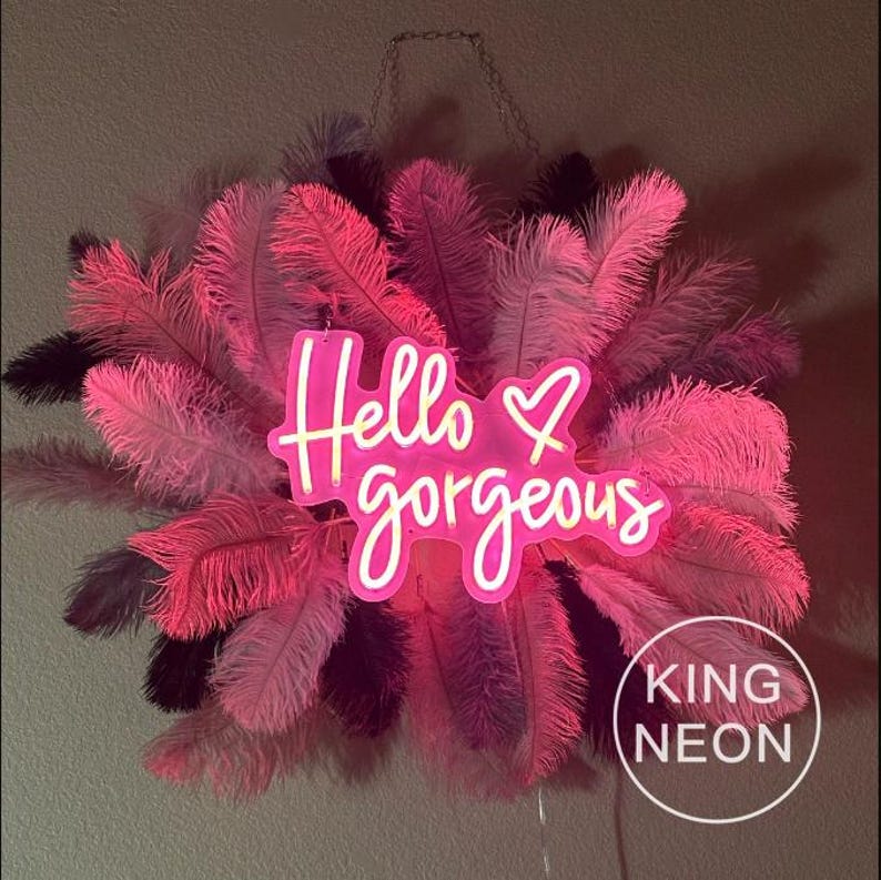 K&ouml;nnte beinhalten: Ein pinkfarbenes Neon-Schild mit dem Text "Hello gorgeous" mit einem Herzsymbol. Das Schild h&auml;ngt an einer Kette und ist von rosa Federn umgeben.