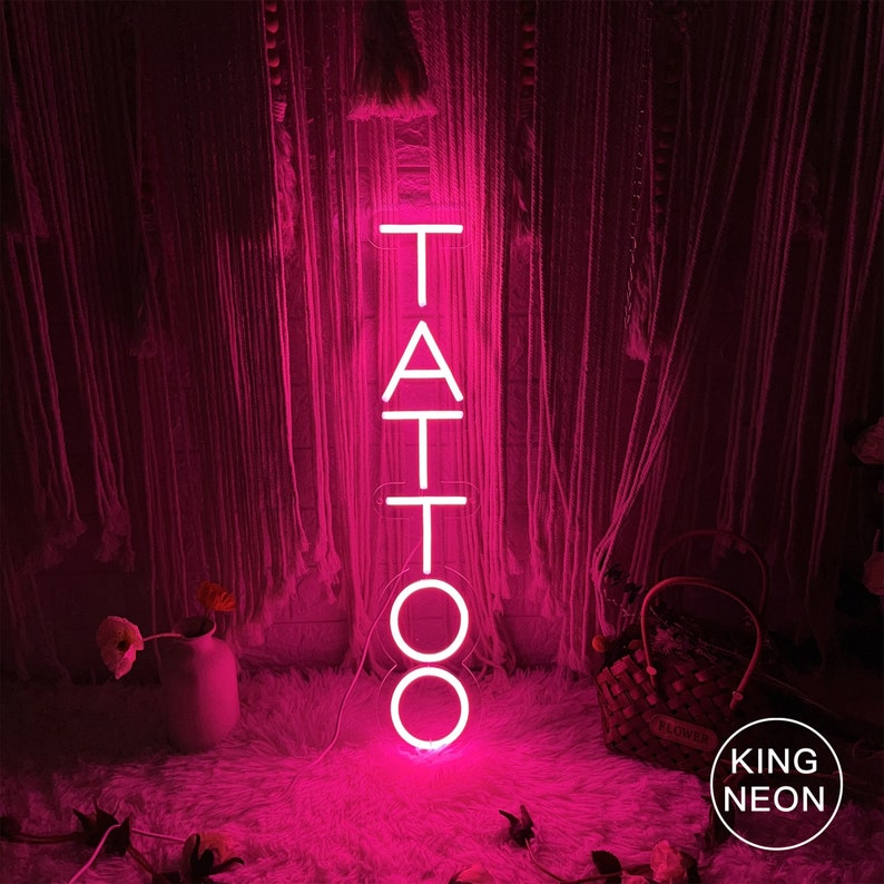 May include: Pink neon sign with the word "TATTOO" in vertical letters. The sign is lit up and is sitting on a pink fluffy surface.