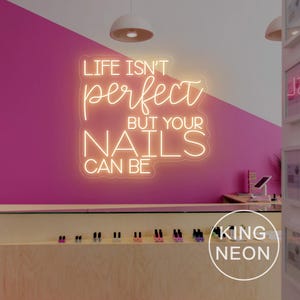 Op de afbeelding: Een neonbord in een nagelsalon met de tekst "LIFE ISN'T perfect BUT YOUR NAILS CAN BE". Het bord gloeit warm op een roze muur. Een display met nagellakflesjes is eronder te zien. King Neon.