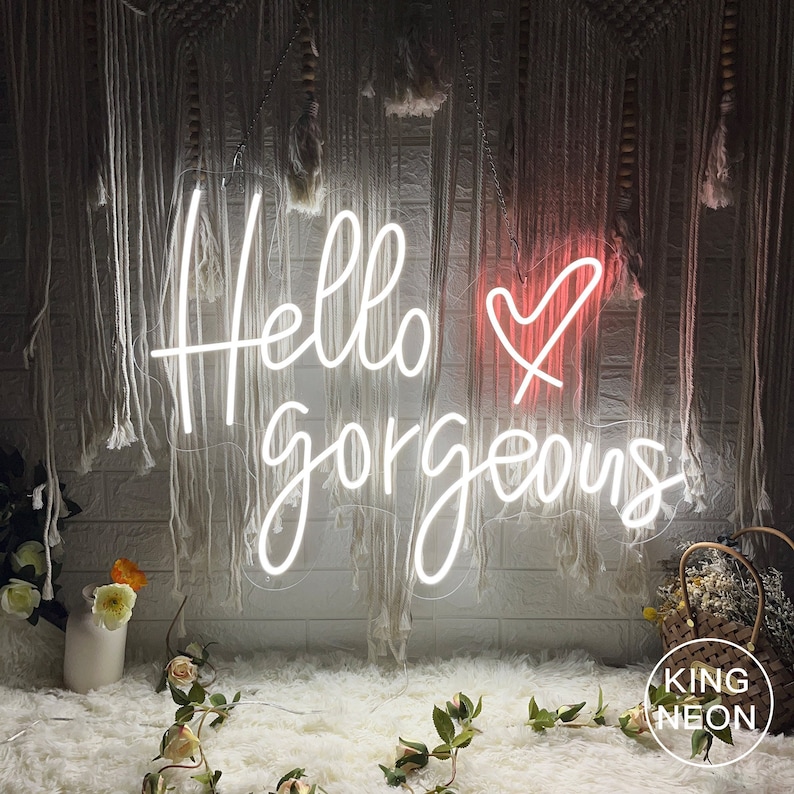 K&ouml;nnte beinhalten: Ein wei&szlig;es Neon-Schild mit dem Text "Hello gorgeous" und einem rosa Herzen. Das Schild h&auml;ngt an einer Wand mit einem wei&szlig;en und grauen, strukturierten Hintergrund.