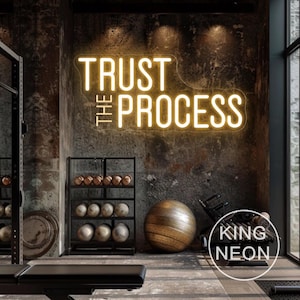 May include: A neon sign with the words "TRUST THE PROCESS" in a gym setting. The sign is illuminated in a warm yellow color and mounted on a textured, dark gray wall. The gym includes exercise equipment, a bench, and a large exercise ball.
