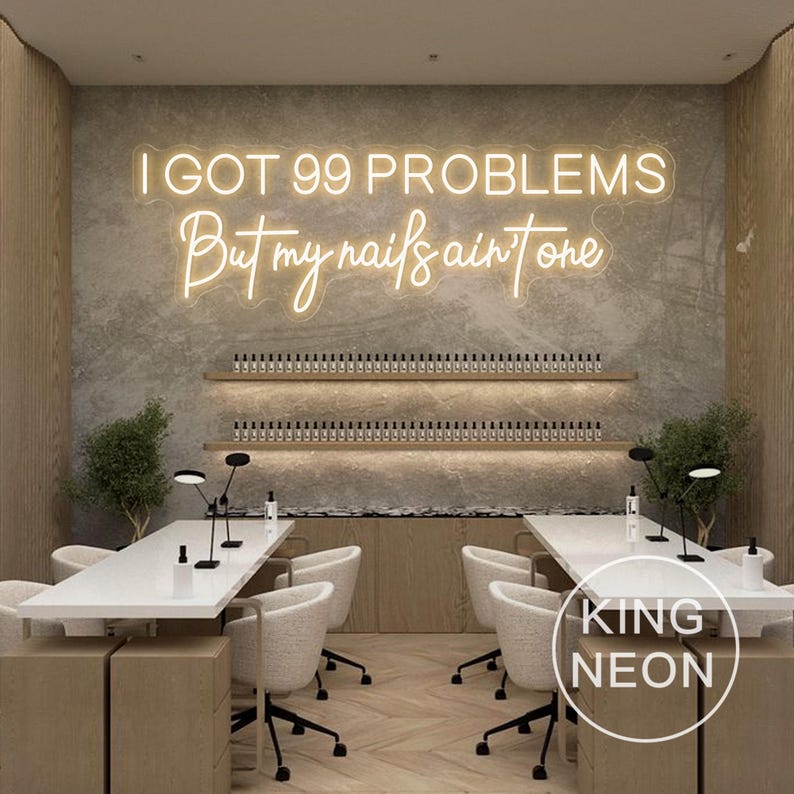 K&ouml;nnte beinhalten: Ein wei&szlig;es Neon-Schild mit dem Text "I got 99 problems but my nails ain't one" h&auml;ngt an einer grauen Wand in einem Nagelstudio. Im Vordergrund stehen zwei wei&szlig;e Manik&uuml;retische mit wei&szlig;en St&uuml;hlen. Das Studio hat ein modernes Design mit Holzakzenten.