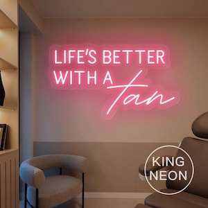 Puede incluir: Un letrero de neón rosa con el texto "Life's Better With A Tan" en una pared blanca. El letrero está iluminado y tiene un contorno blanco. El texto "King Neon" es visible en la esquina inferior derecha de la imagen.