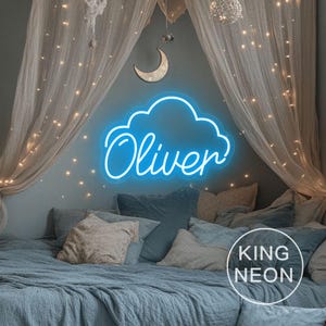 Pode incluir: Uma placa de néon azul em forma de nuvem com o nome "Oliver" em escrita branca. A placa está montada numa parede cinzenta acima de uma cama com roupa de cama e almofadas azuis. O quarto é decorado com luzes de fadas e uma lua crescente.