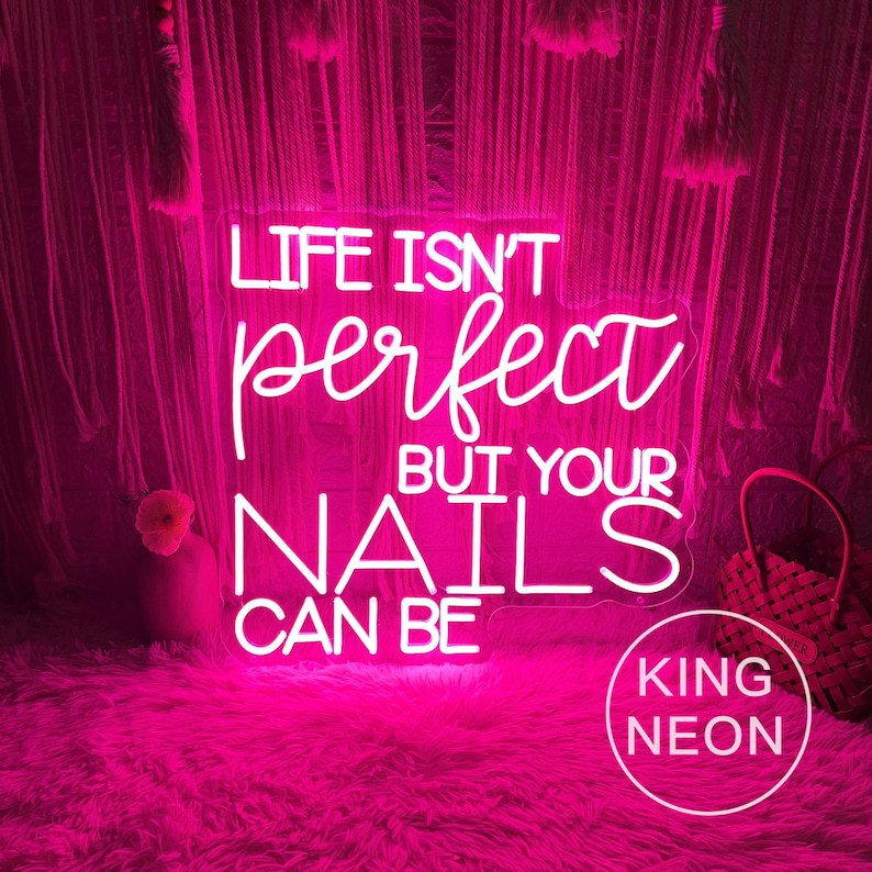 Op de afbeelding: Een helder roze neonbord met de tekst "LIFE ISN'T perfect BUT YOUR NAILS CAN BE". Het bord wordt omgeven door een roze, pluizig tapijt en een decoratieve achtergrond. Het bord heeft ook de woorden "KING NEON" in een cirkel.